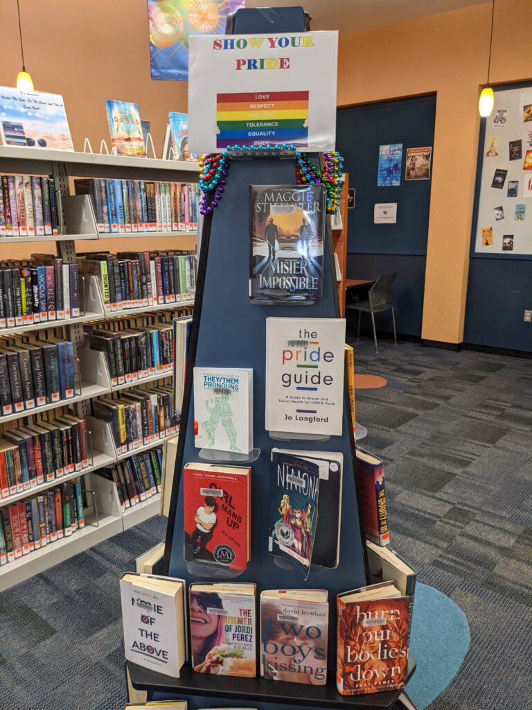 A Pride Month display in a public library. It is freestanding, round, with a sign on top that says "Show Your Pride."